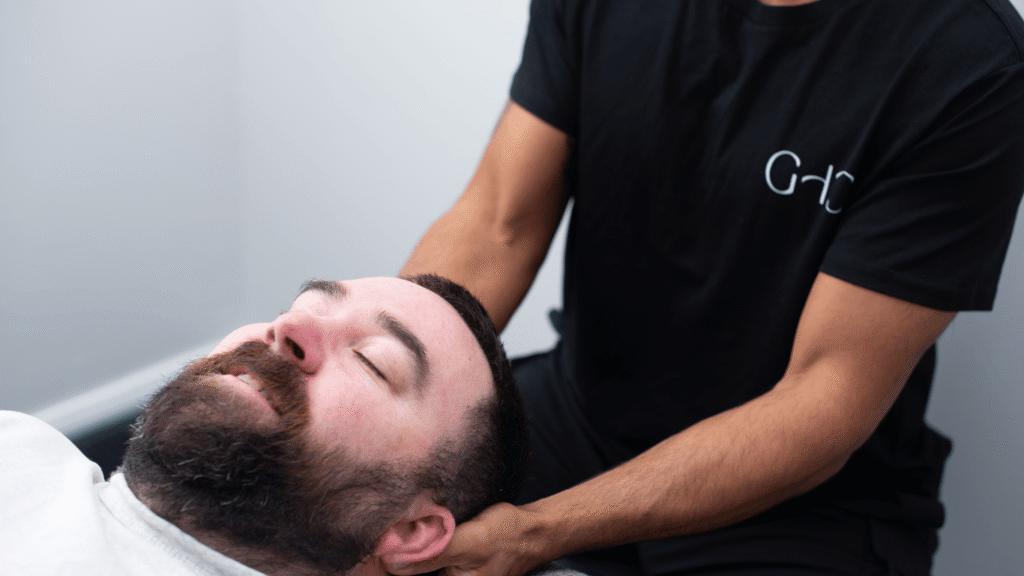 A man receives a neck massage from another person in a black t-shirt in a bright room.