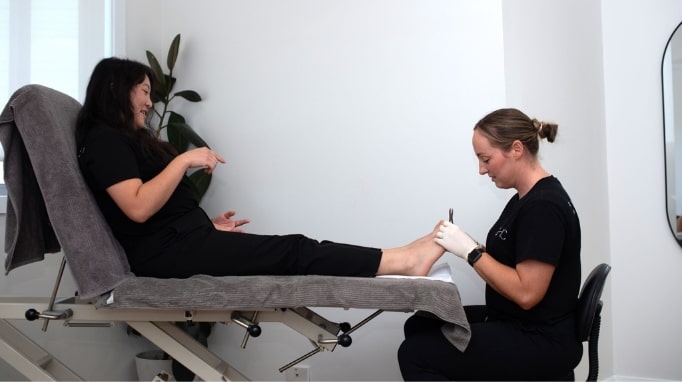 Two women in a clinic; one receives a foot treatment from a professional wearing gloves.