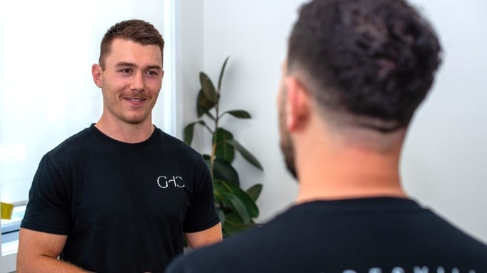 Two men stand and talk indoors, one facing the camera and one with his back to the camera.