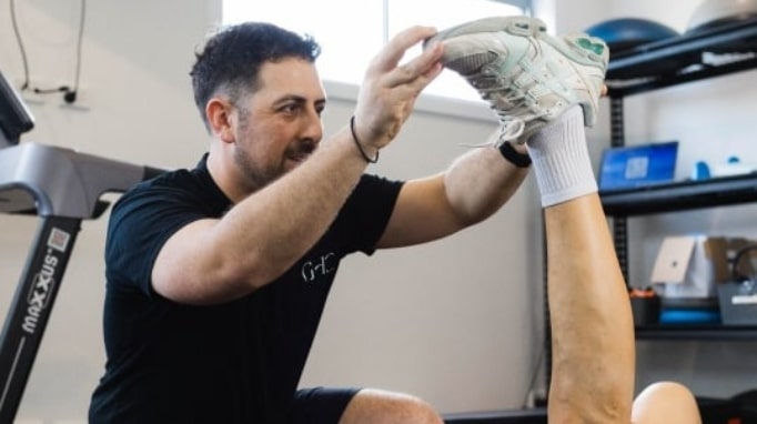 Trainer assists person stretching their leg in a gym setting.