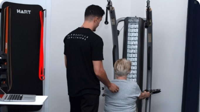 A man assists an older woman using a weight machine in a fitness or rehabilitation setting.