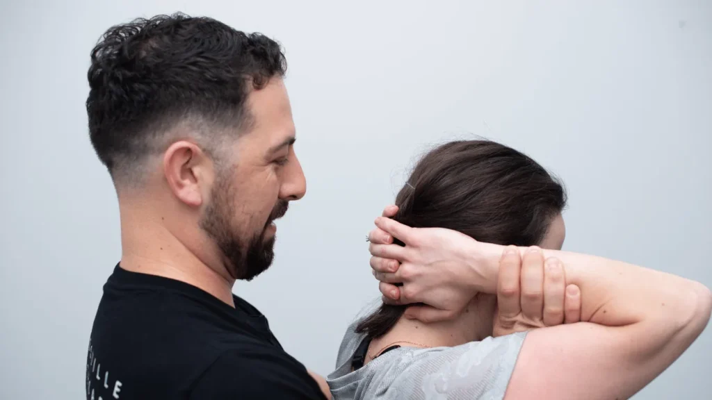 A Gladesville chiropractor assists a woman stretching her neck, hands clasped behind her head.