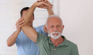 Older man receives physical therapy for lower back pain relief, therapist assists with arm movement.